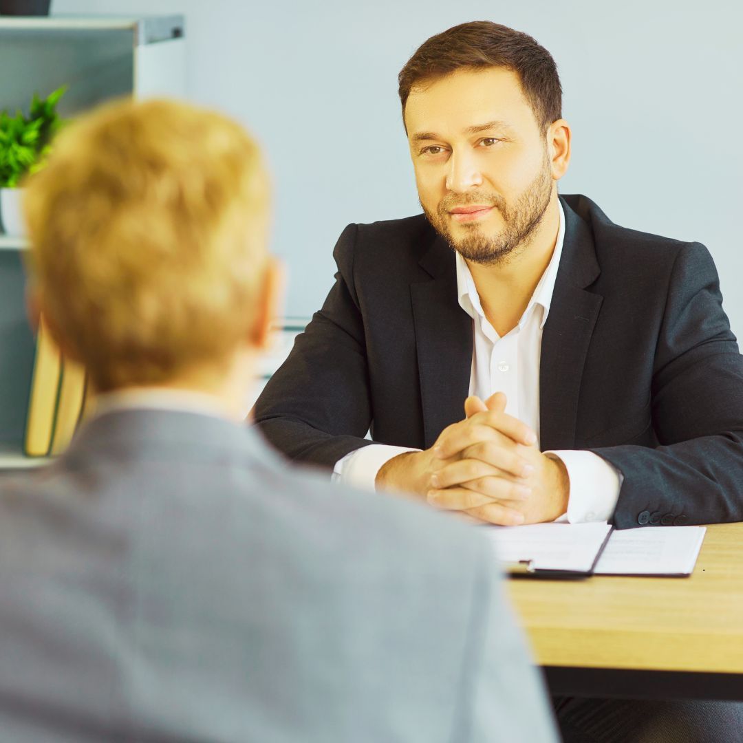 Man in a suit interviewing another person at a desk. Both are in suits.