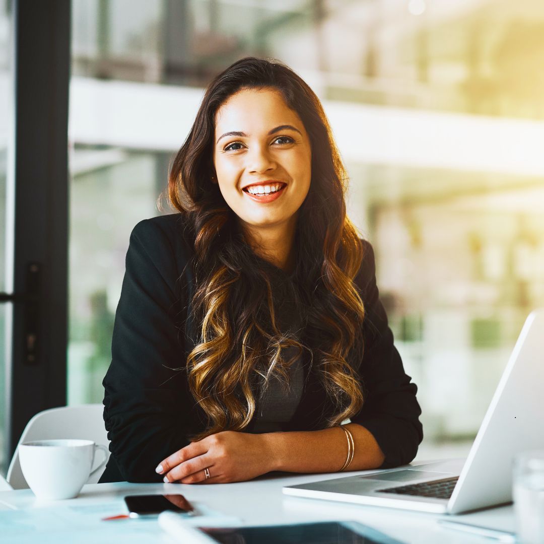 Woman with long dark hair smiles while seated at a desk with a laptop, coffee cup, and tablet.
