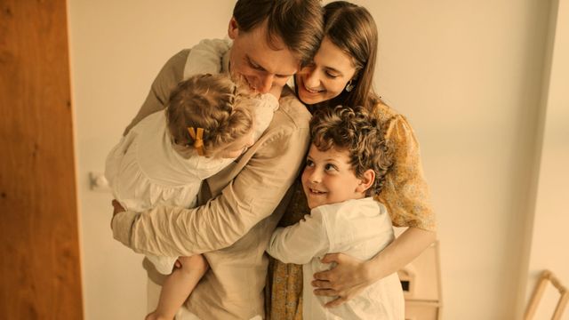 Family of four embracing, smiling. Beige and white clothing, warm lighting.