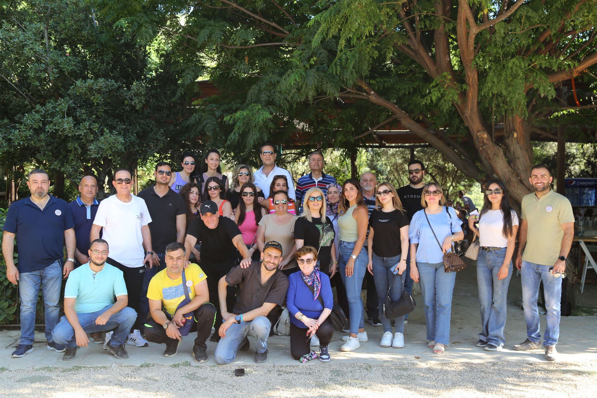 Group of people posing outdoors under trees. Some are kneeling, smiling. Sunny day.