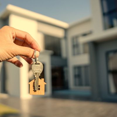 Hand holding keys with house-shaped keychain in front of a modern house.
