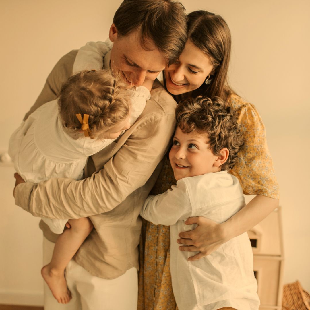 Family of four embraces indoors; smiles and affection.