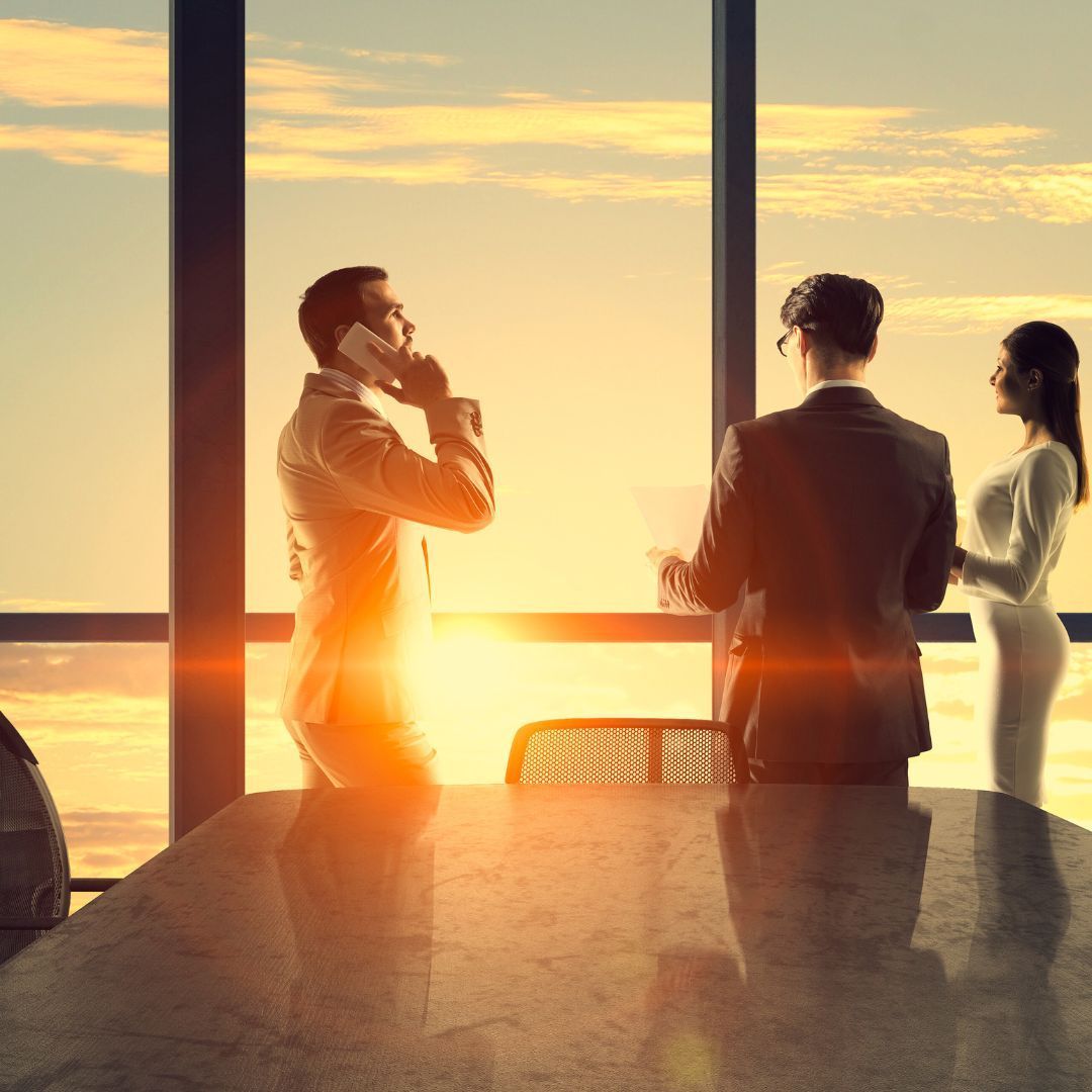 Businesspeople in office, near large windows, illuminated by sunset. One on the phone.