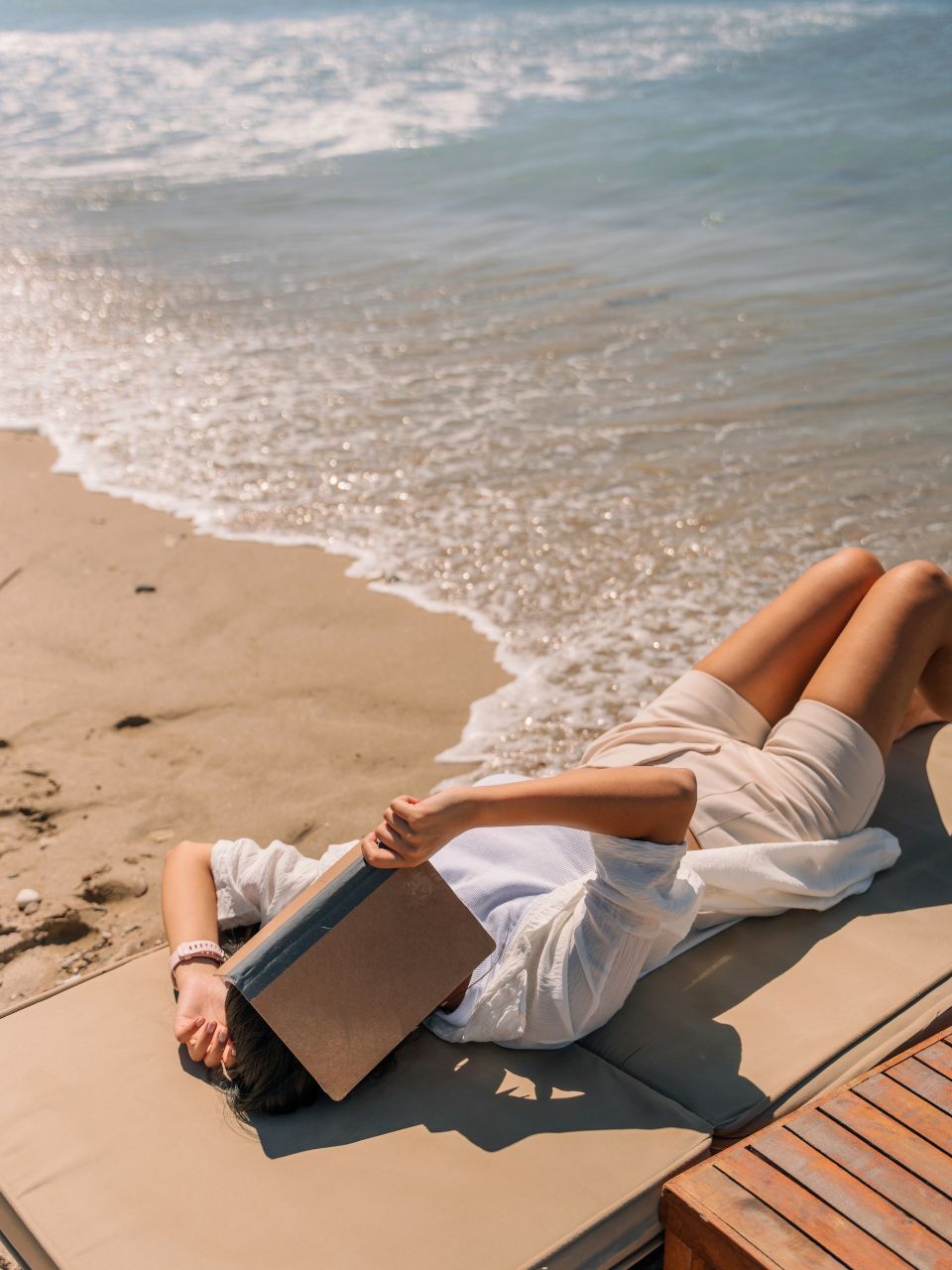 Person lying on a beach chair, covering face with a book, ocean in the background.