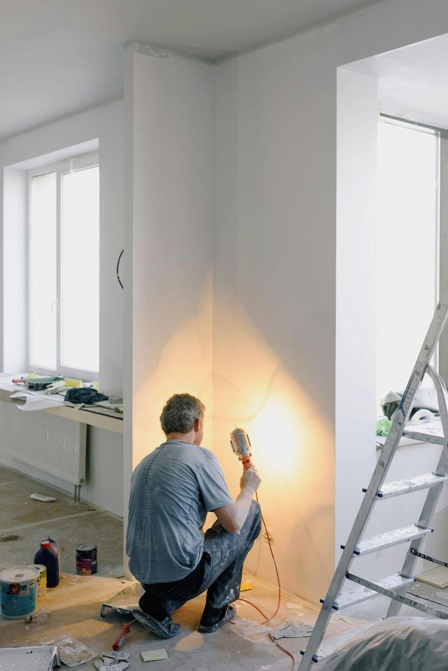 Person painting a white wall indoors beside a ladder, with tools on the floor.