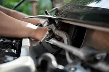 Hands working on a car engine in a garage, with tools and metal parts visible.