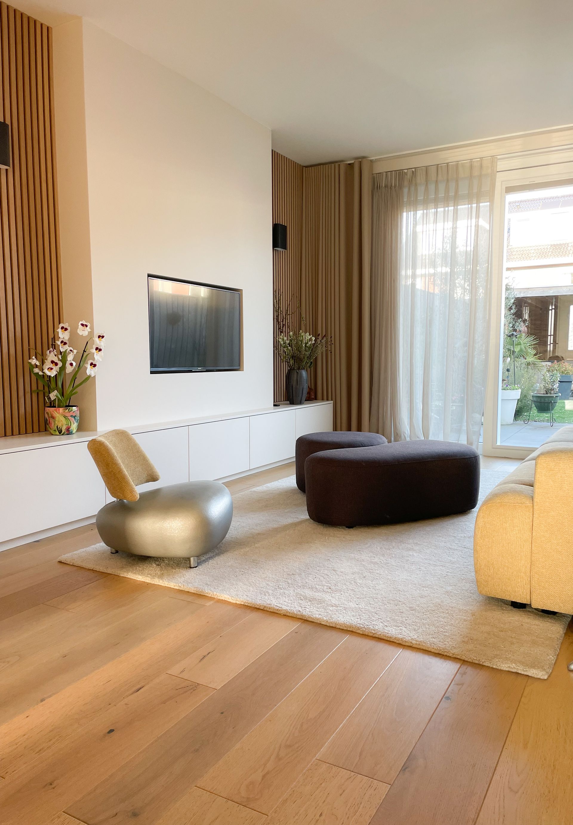 Een minimalistisch ingerichte woonkamer met een geel/groene bank voor een op maat gemaakt en ontworpen wandmeubel waar de televisie in geïntegreerd is. Naast het wandmeubel staat een fauteuil van Leolux waarvoor de rugleuning een nieuwe stof gekregen heeft zodat deze weer in het interieur past.  