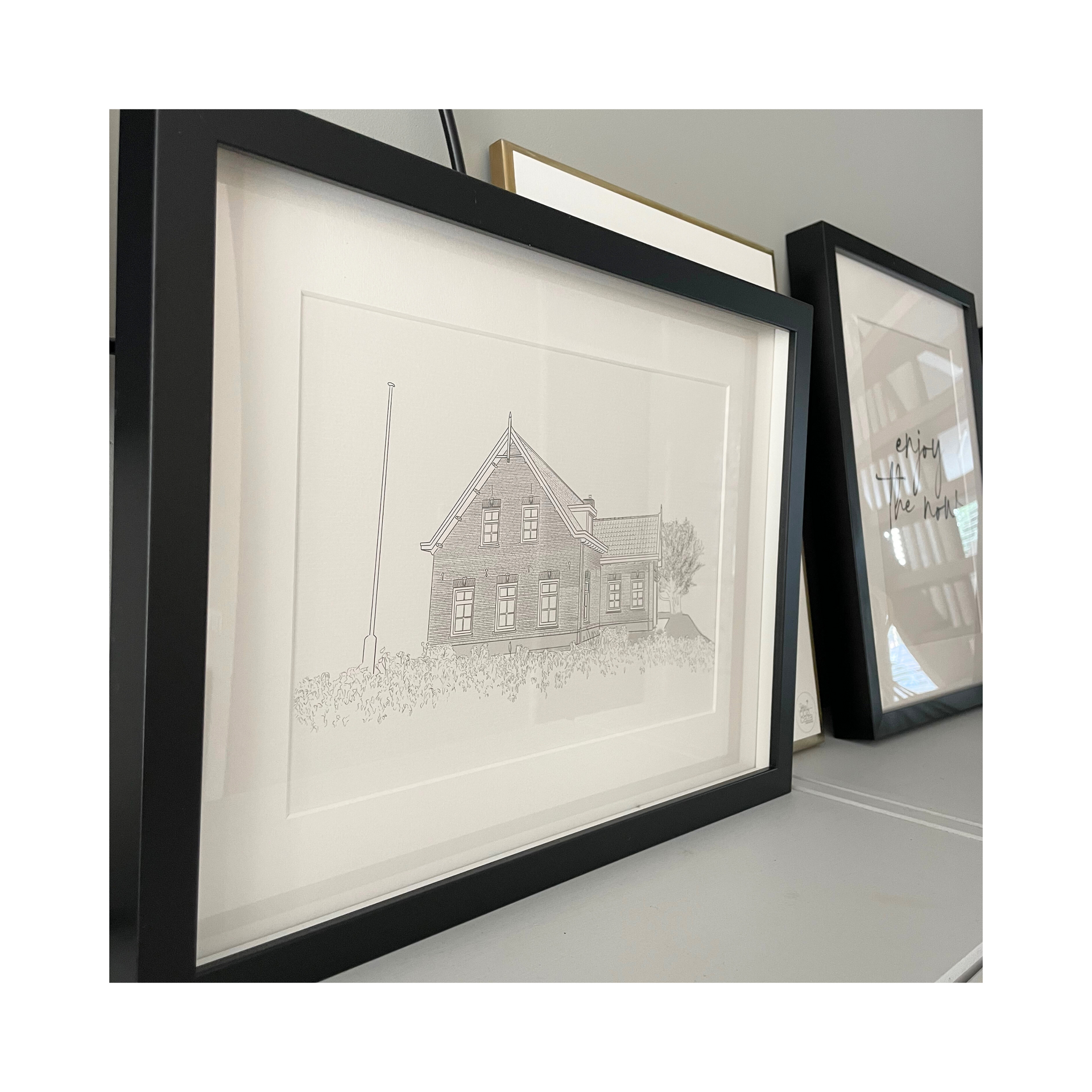 Twee ingelijste tekeningen van een huis staan ​​op een plank.