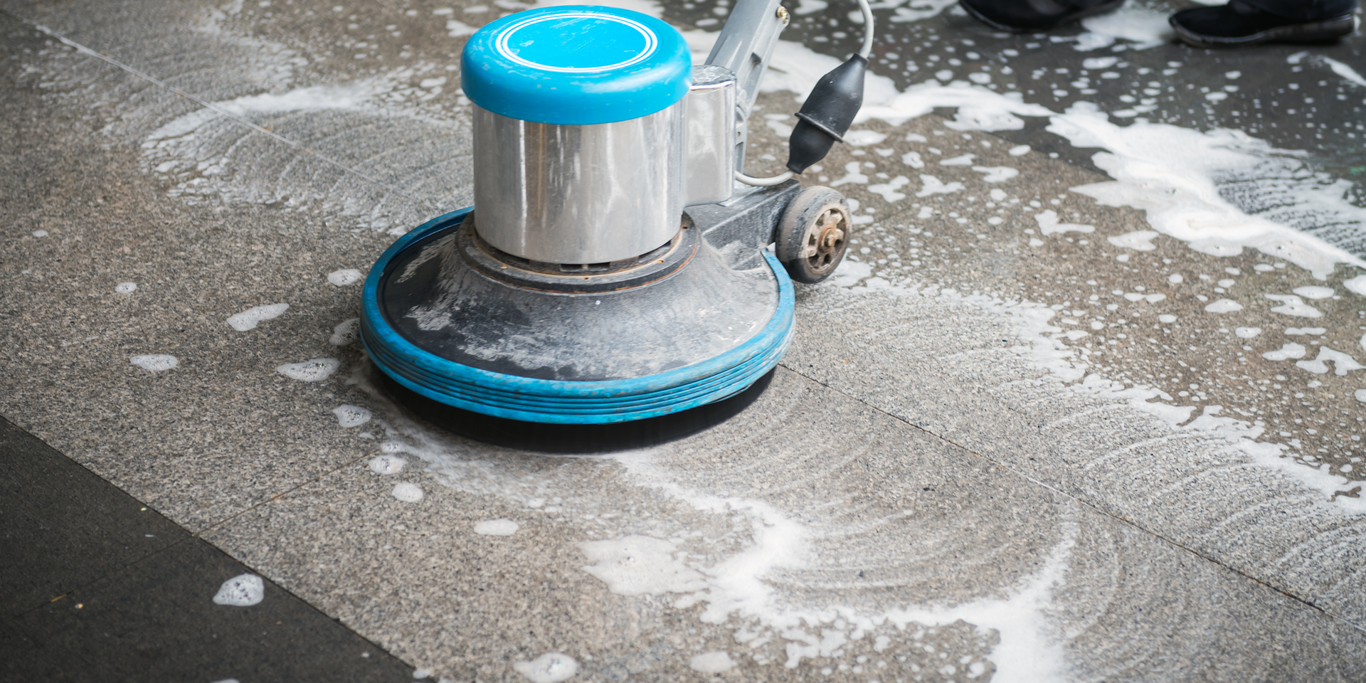 A floor cleaning machine in action, scrubbing a concrete surface with foamy detergent.