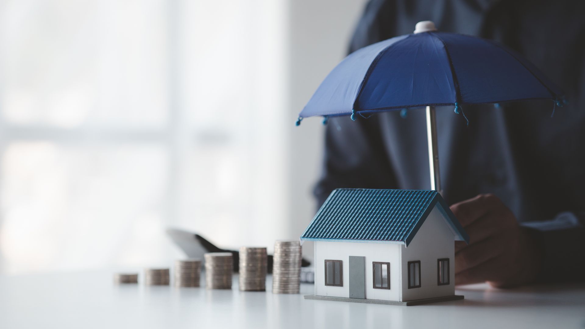 A person is holding an umbrella over a model house and coins.