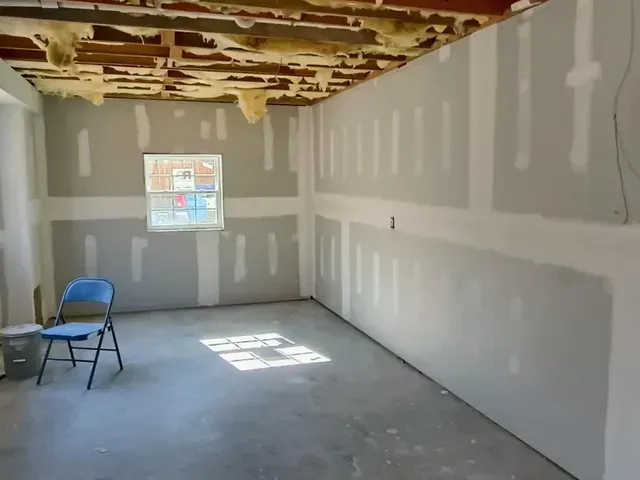 Empty unfinished room with drywall patches, exposed ceiling, a blue chair, and a sunlit window.