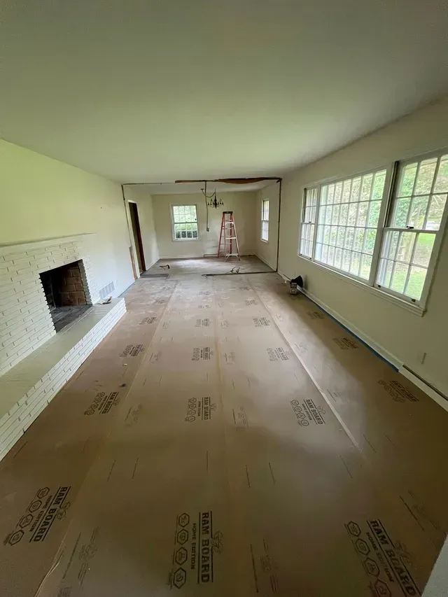 Long, empty living room under renovation with covered floors, fireplace, windows, and a chandelier