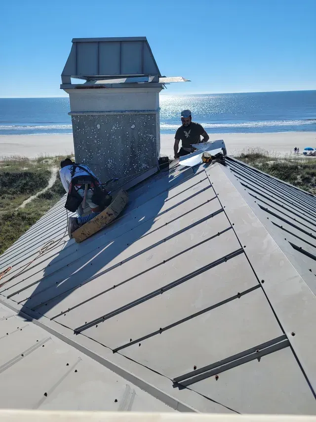 Two workers on a metal rooftop near a beach, working beside a chimney under a bright blue sky