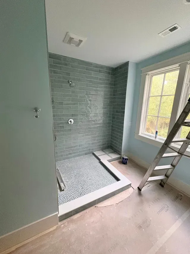 Small tiled shower under renovation with teal walls, window light, and a ladder nearby