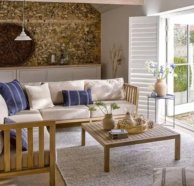 Cozy living room with beige sofa, blue pillows, wooden coffee table, woven rug, and open shutters to bright garden view