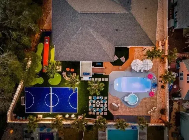 Aerial view of a backyard with a pool, basketball court, patio seating, and a house roof.