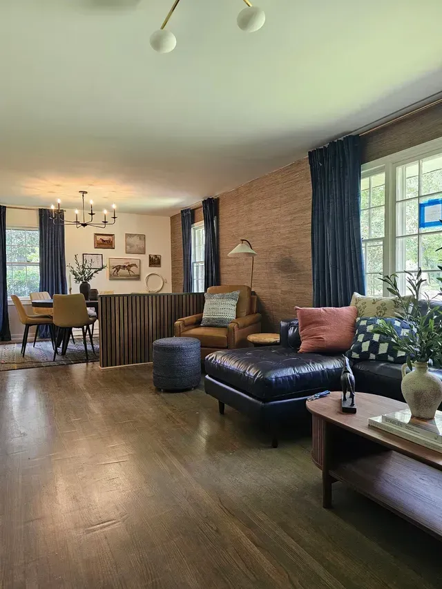 Cozy living room with dark sofa, armchairs, wood floors, and large windows with dark curtains