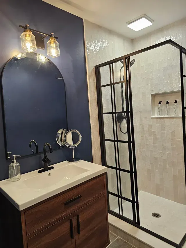 Modern bathroom with black-framed glass shower, navy accent wall, and wooden vanity with gold sconces