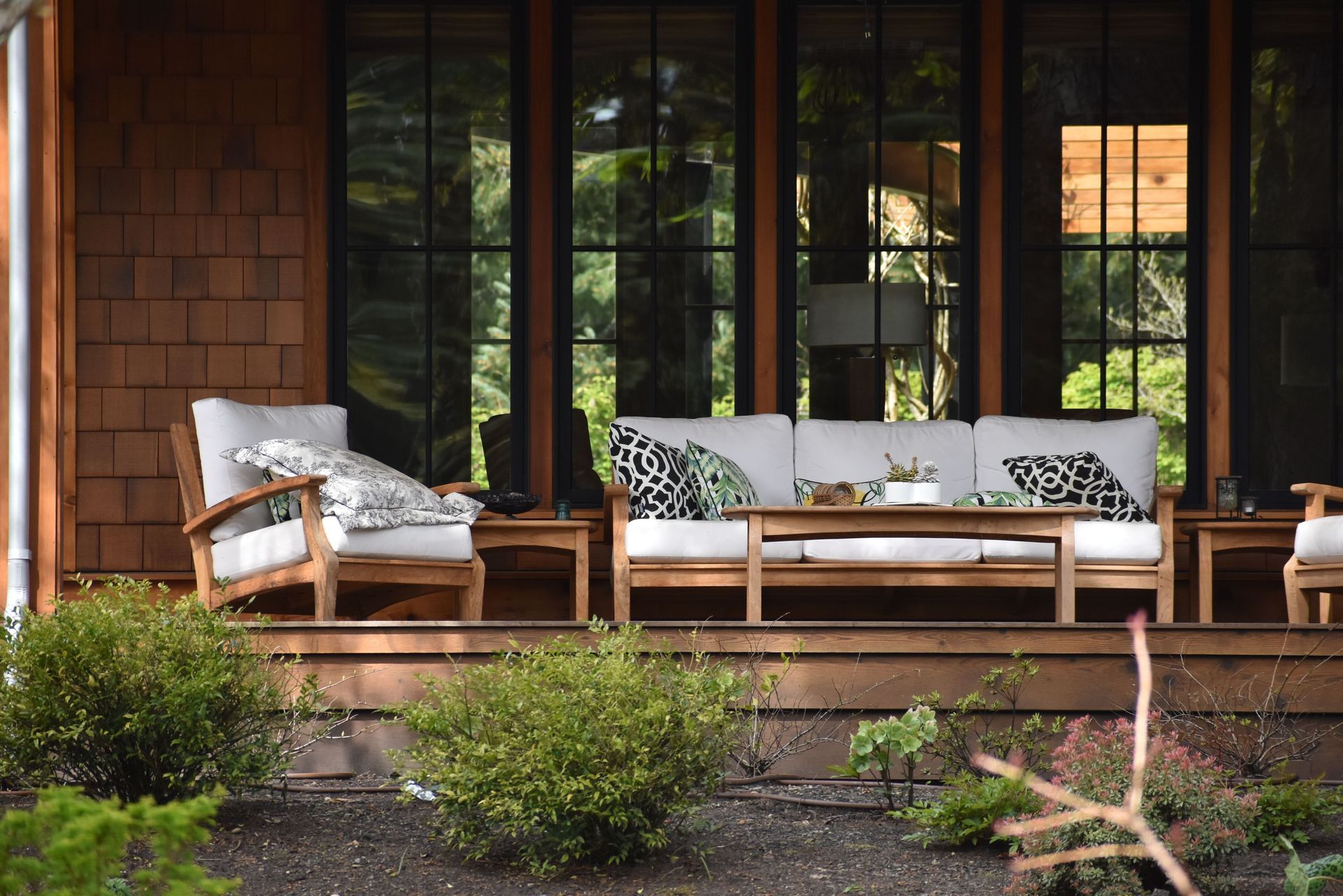 Wooden porch with white cushioned chairs and bench, framed by dark glass doors and greenery in front
