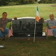 Two men are sitting in front of a grave with flags.