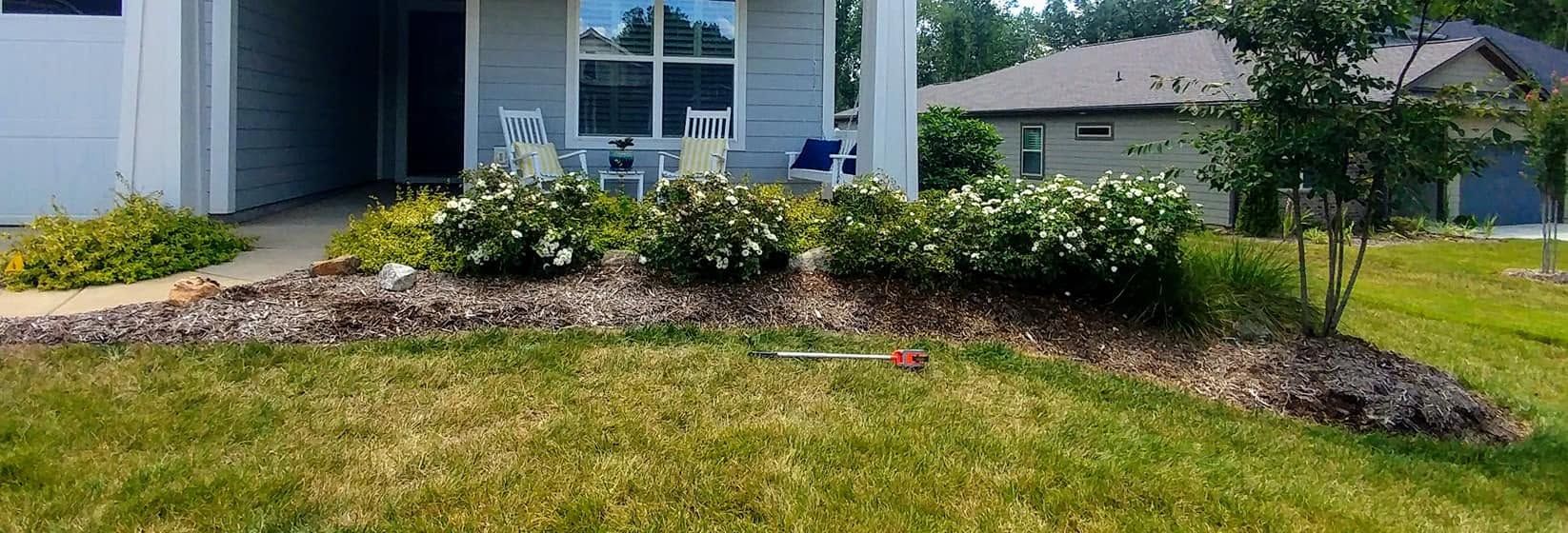 Front yard flower bed with shrubs and mulch beside a house, bordered by green lawn and driveway