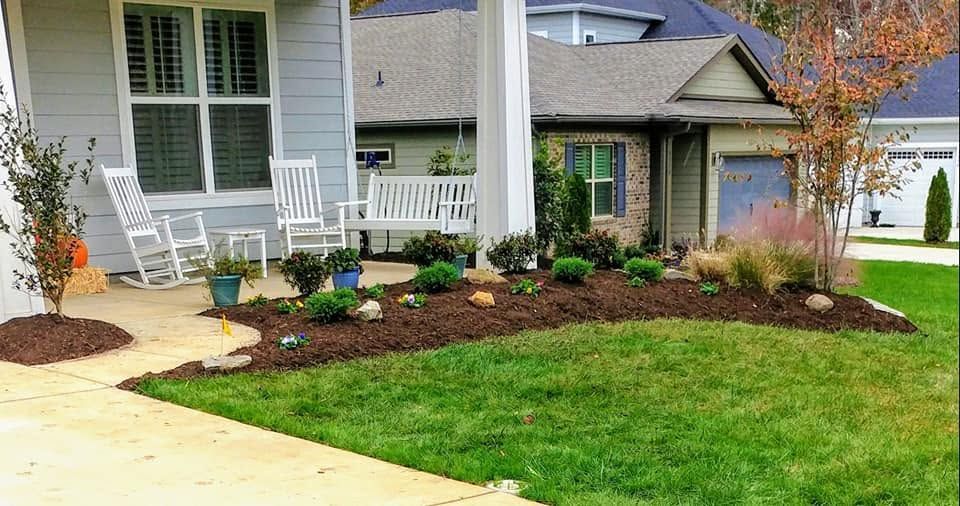 Front yard landscaping with fresh mulch, shrubs, flowers, and a green lawn beside a porch and driveway