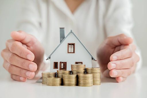 A person is holding a model house and stacks of coins.