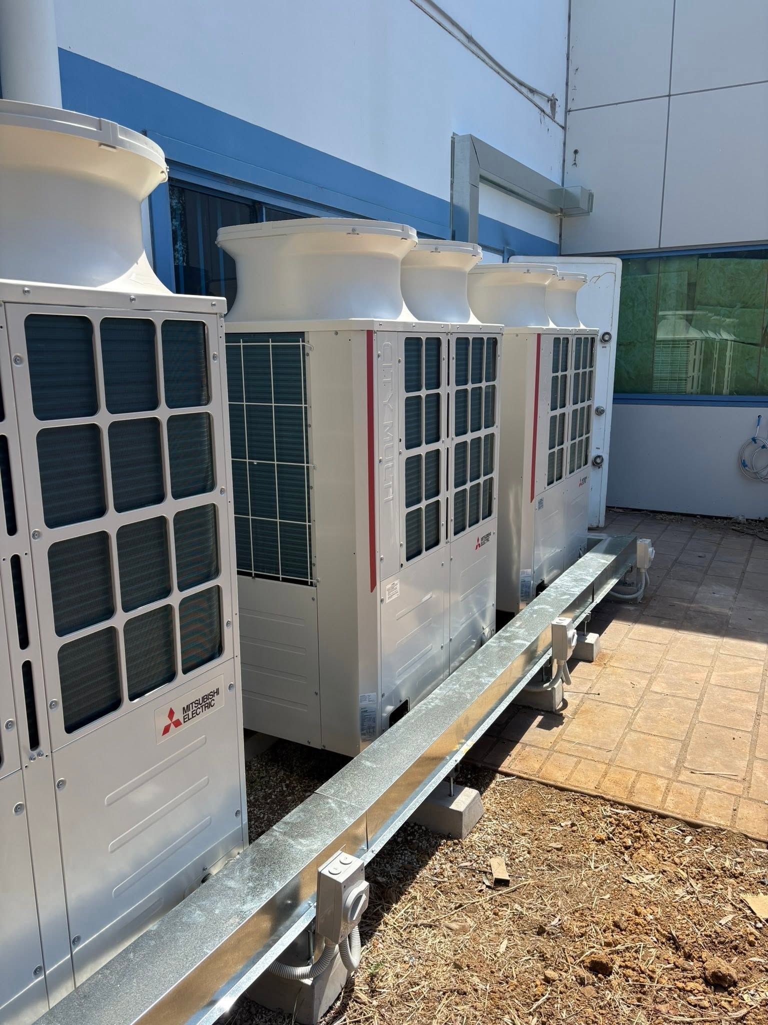 Row of outdoor HVAC units beside a white building, with blue trim and a gravel walkway.