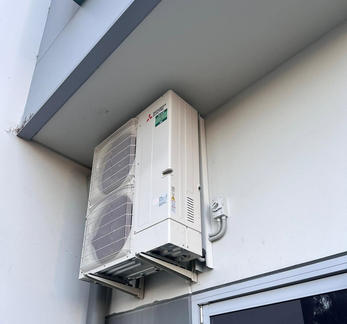 Wall-mounted outdoor air conditioner unit beneath a building overhang, with a window below.
