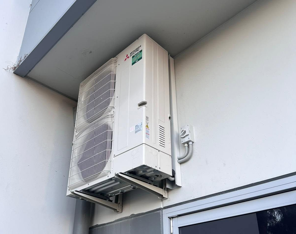 Wall-mounted outdoor air conditioner unit beneath an overhang, with a white exterior and visible vents.