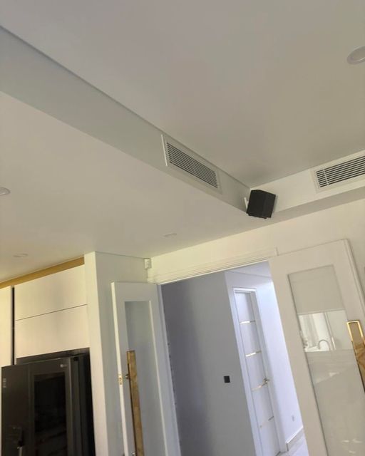 Modern white interior with ceiling vents, recessed lights, and a doorway near a black wall-mounted speaker.