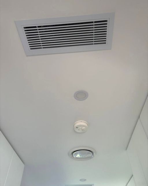 Ceiling with a rectangular air vent, smoke detector, and recessed light in a white hallway