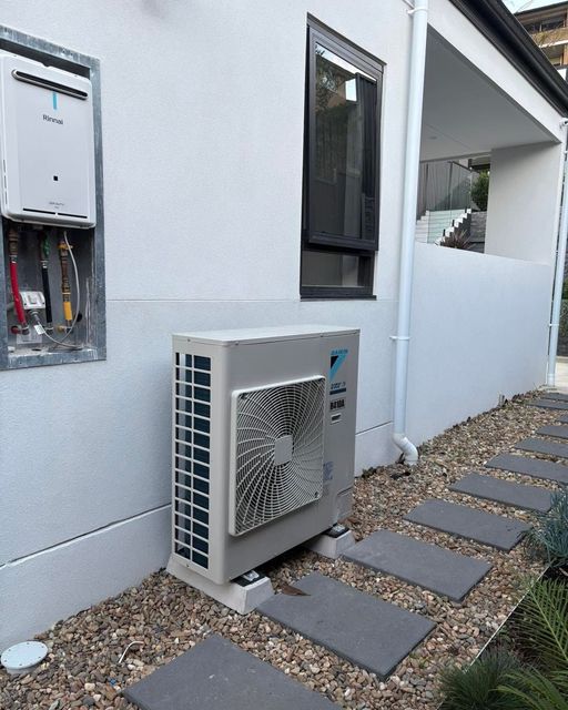 Outdoor air conditioner unit beside a white house wall and stepping stones in a gravel yard