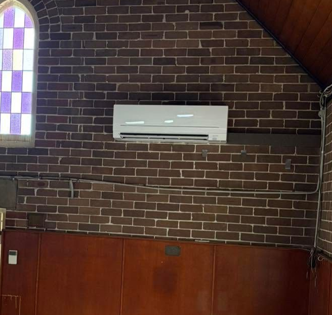 White wall-mounted air conditioner on a dark brick church wall above wood paneling and stained glass window