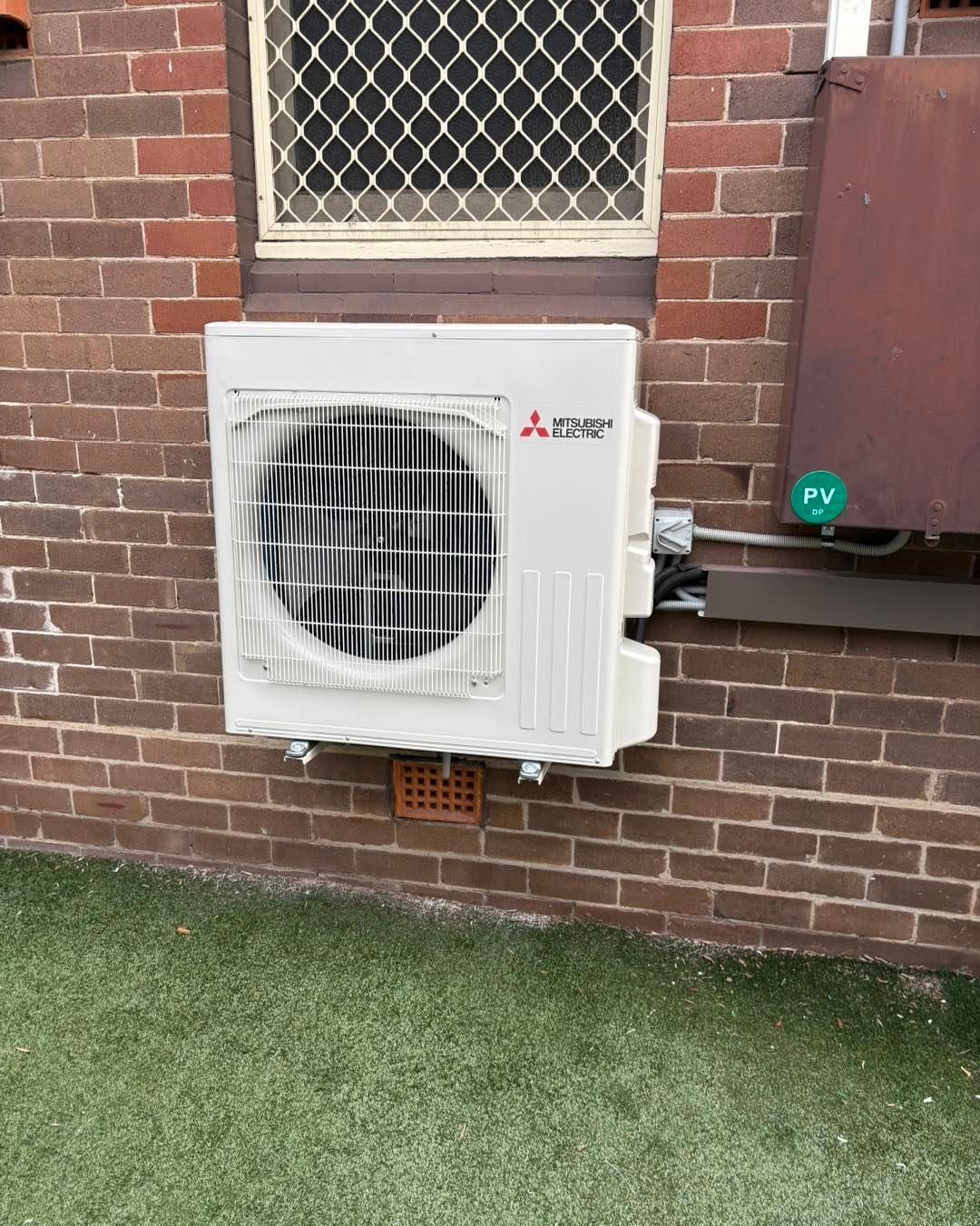 Outdoor wall-mounted Mitsubishi air conditioner unit on a brick building with grass below.