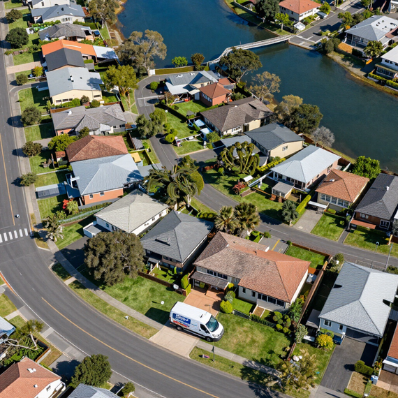 Aerial view of suburban homes along a curved road beside a canal or lake, with parked vehicles and trees.