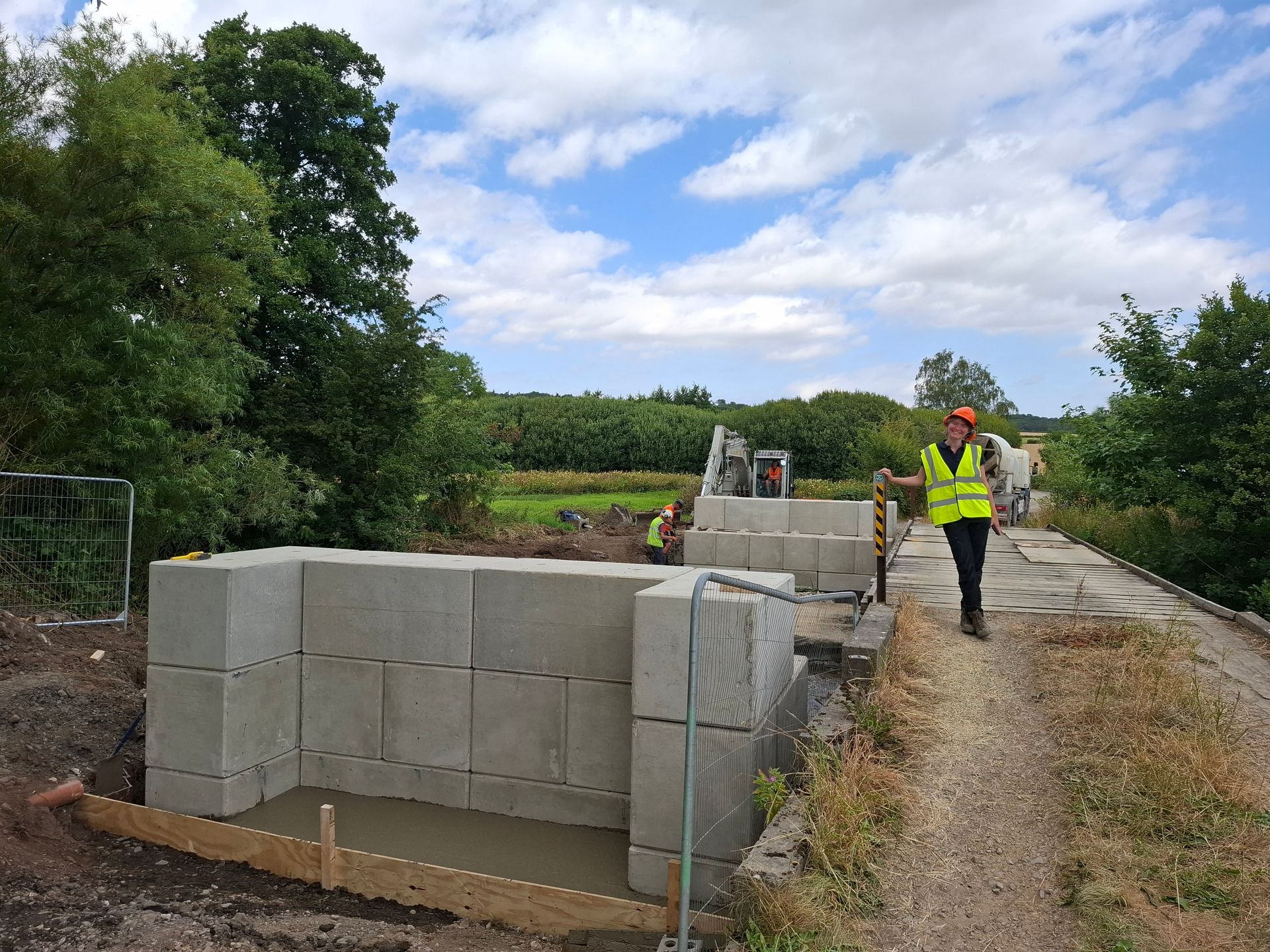 woman stood by a bridge that is being built over a stream wearing a hard hat and a high visibility jacket.