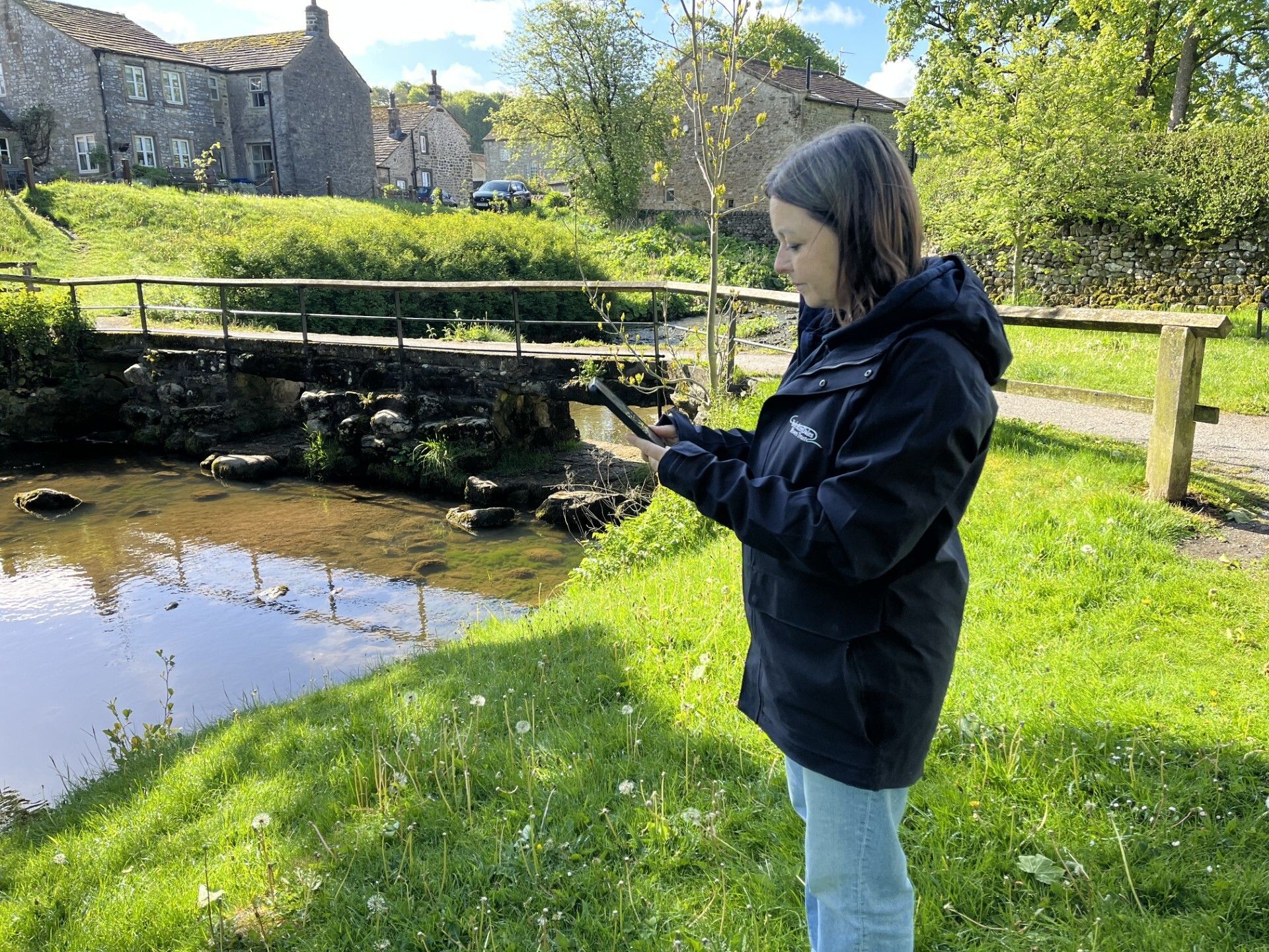 A woman stood by a stream taking notes and looking at a small stone ford in the stream.
