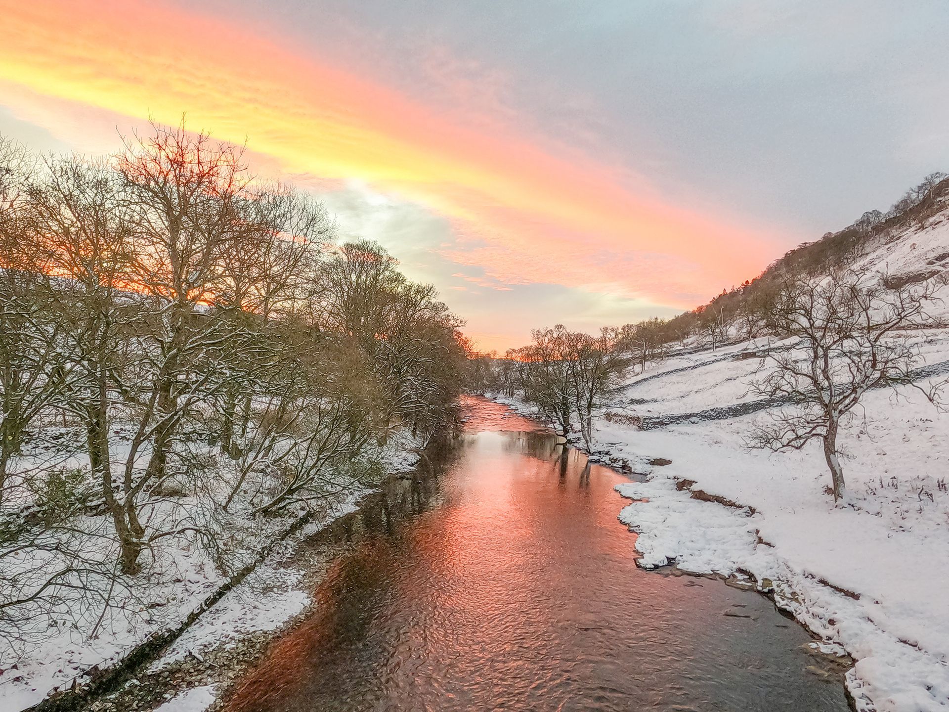 A winter river flanked by bare trees and a pink sunrise