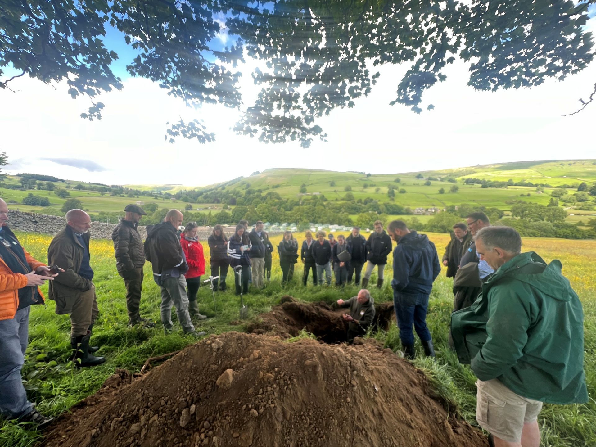 Man stood in a hole in a field surrounded by an audience and talking about soil.