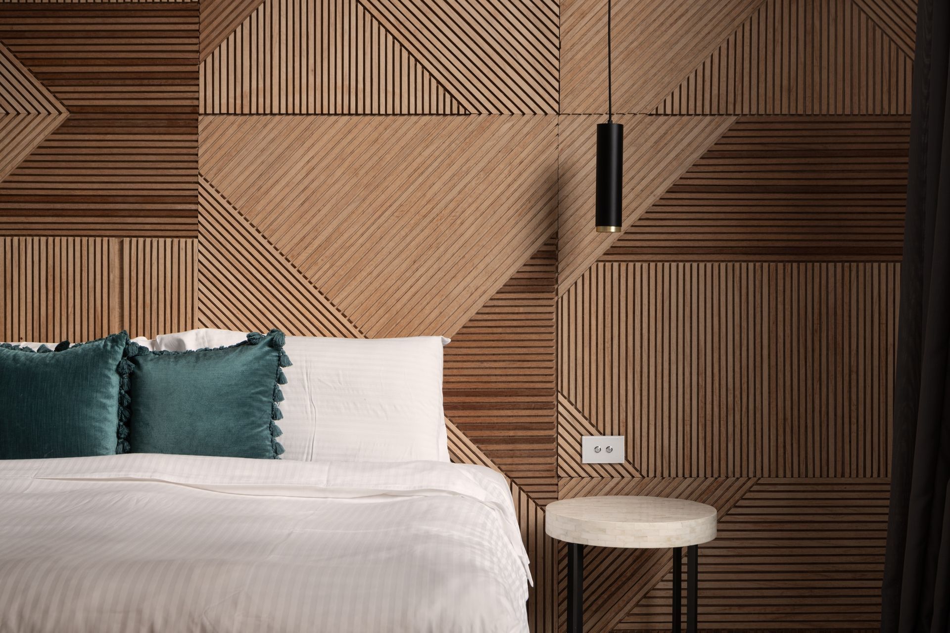 Modern bedroom with geometric wood wall, white bed with teal pillows, and a small bedside table and lamp