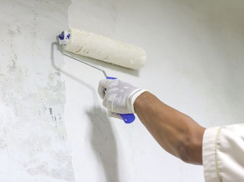 Person painting a white wall with a paint roller, wearing a white glove and blue-handled tool