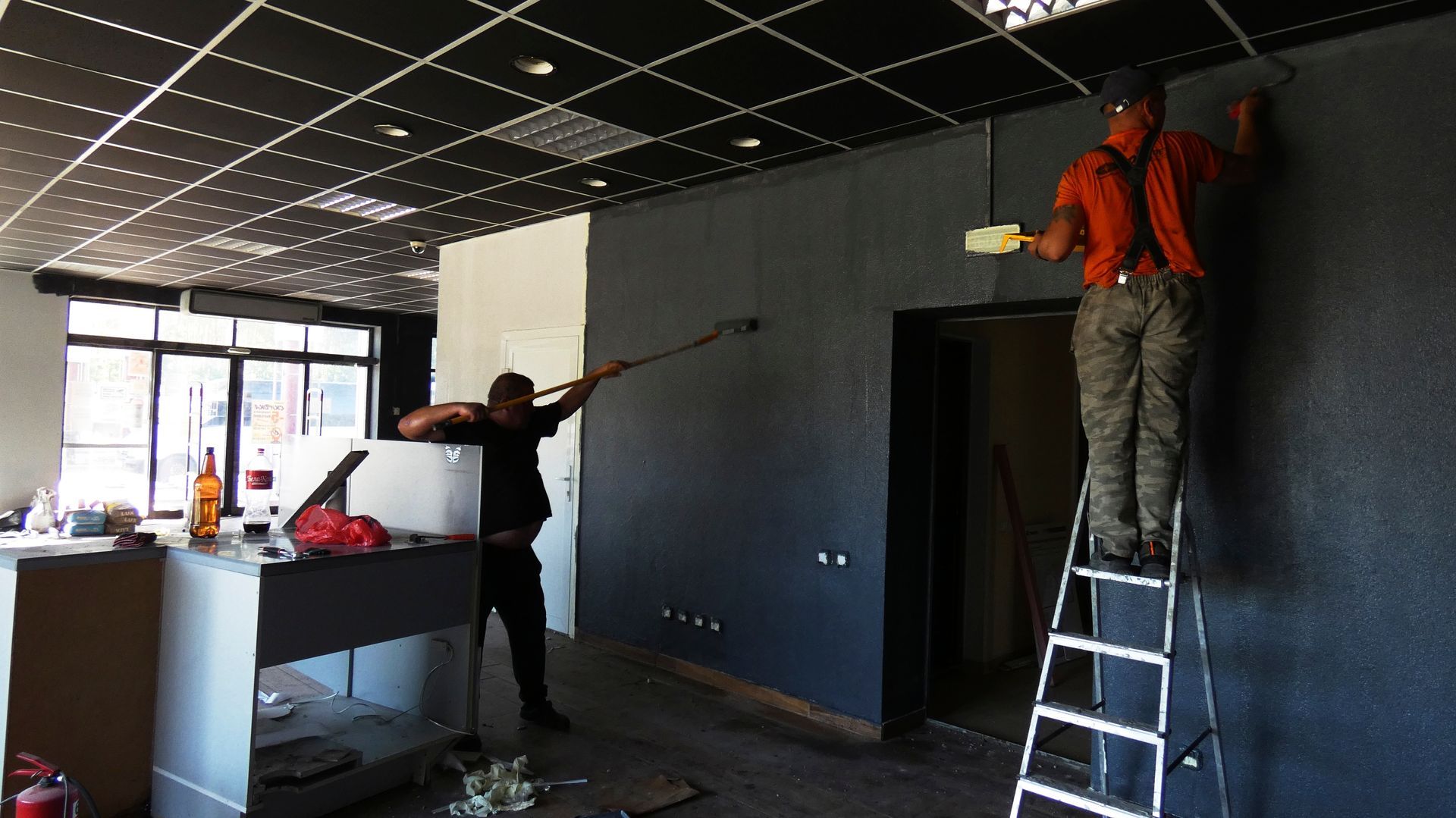 Two workers installing a suspended ceiling in a dark room, one on a ladder and one on the floor.