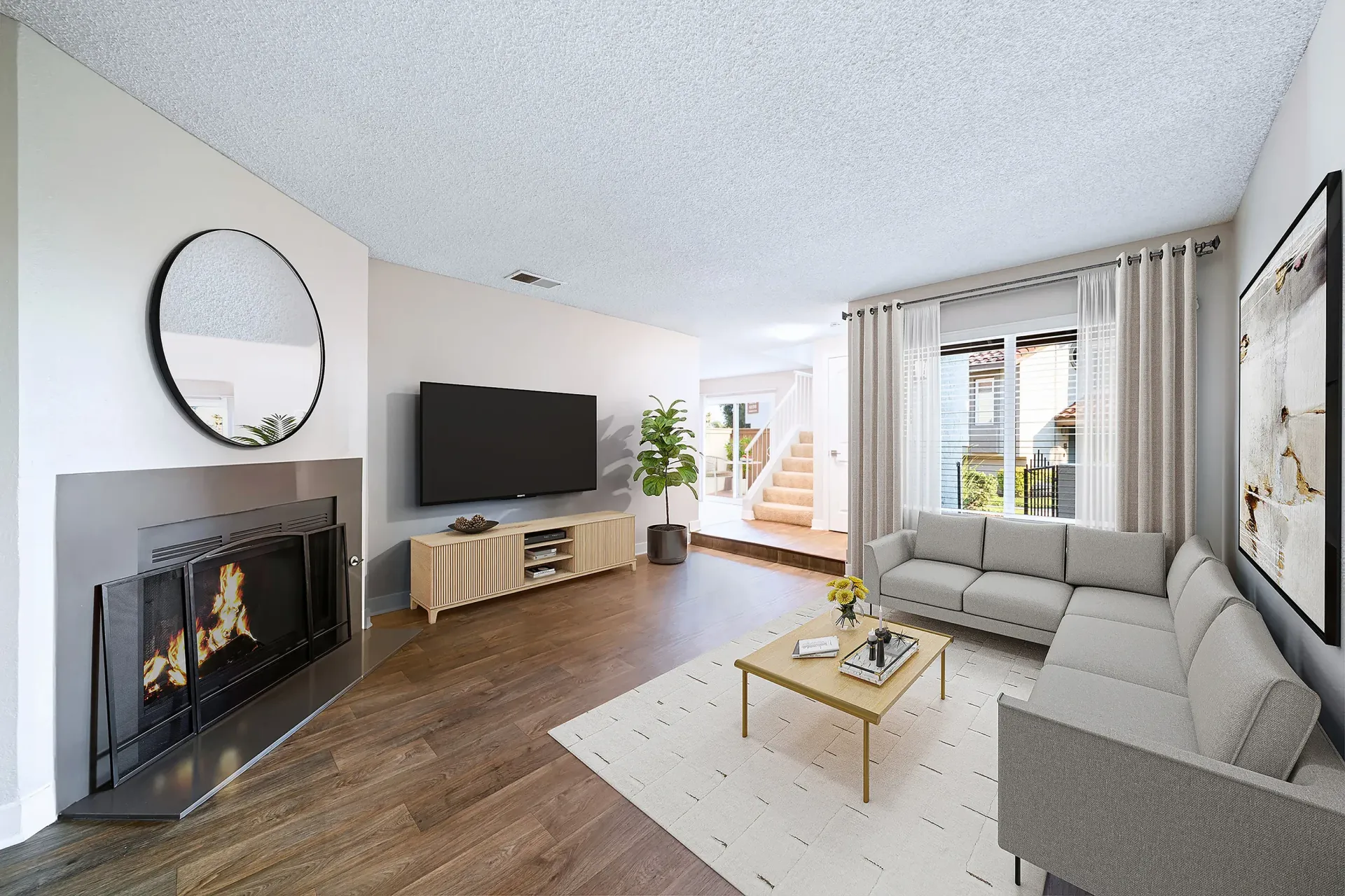 Living room with a gray L-shaped sofa, a fireplace, a wall-mounted TV, and a large window with sheer curtains at Sonora at Alta Loma apartments in Alta Loma, CA.