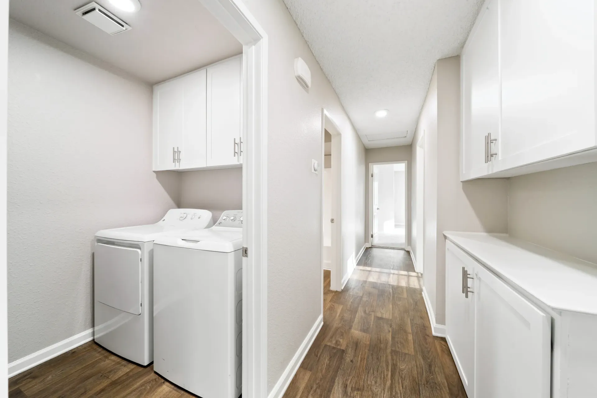 Laundry closet with a white washer and dryer, white cabinets, and a hallway with wood-look flooring at Sonora at Alta Loma apartments in Alta Loma, CA.