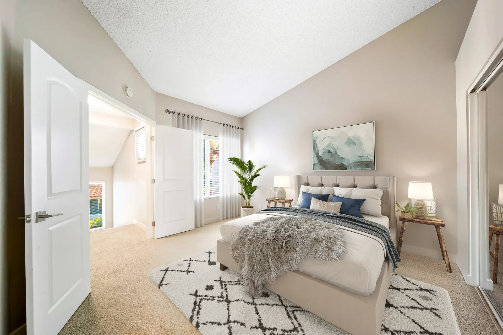 Bedroom with beige walls, tufted upholstered bed, two nightstands, plant, and white curtains at Sonora at Alta Loma apartments in Alta Loma, CA.