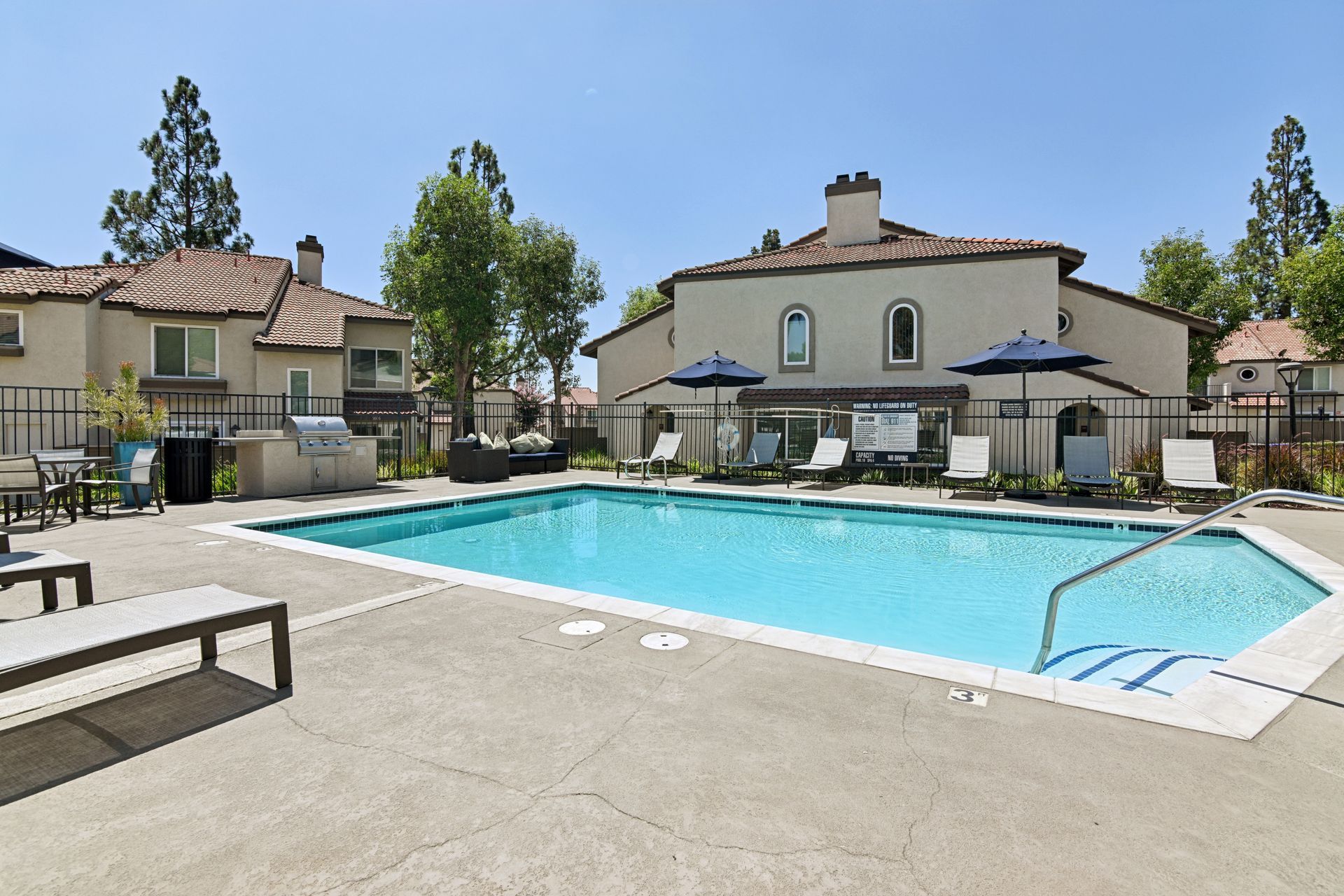 Outdoor apartment pool courtyard with turquoise water, lounge chairs, and beige buildings under a clear sky