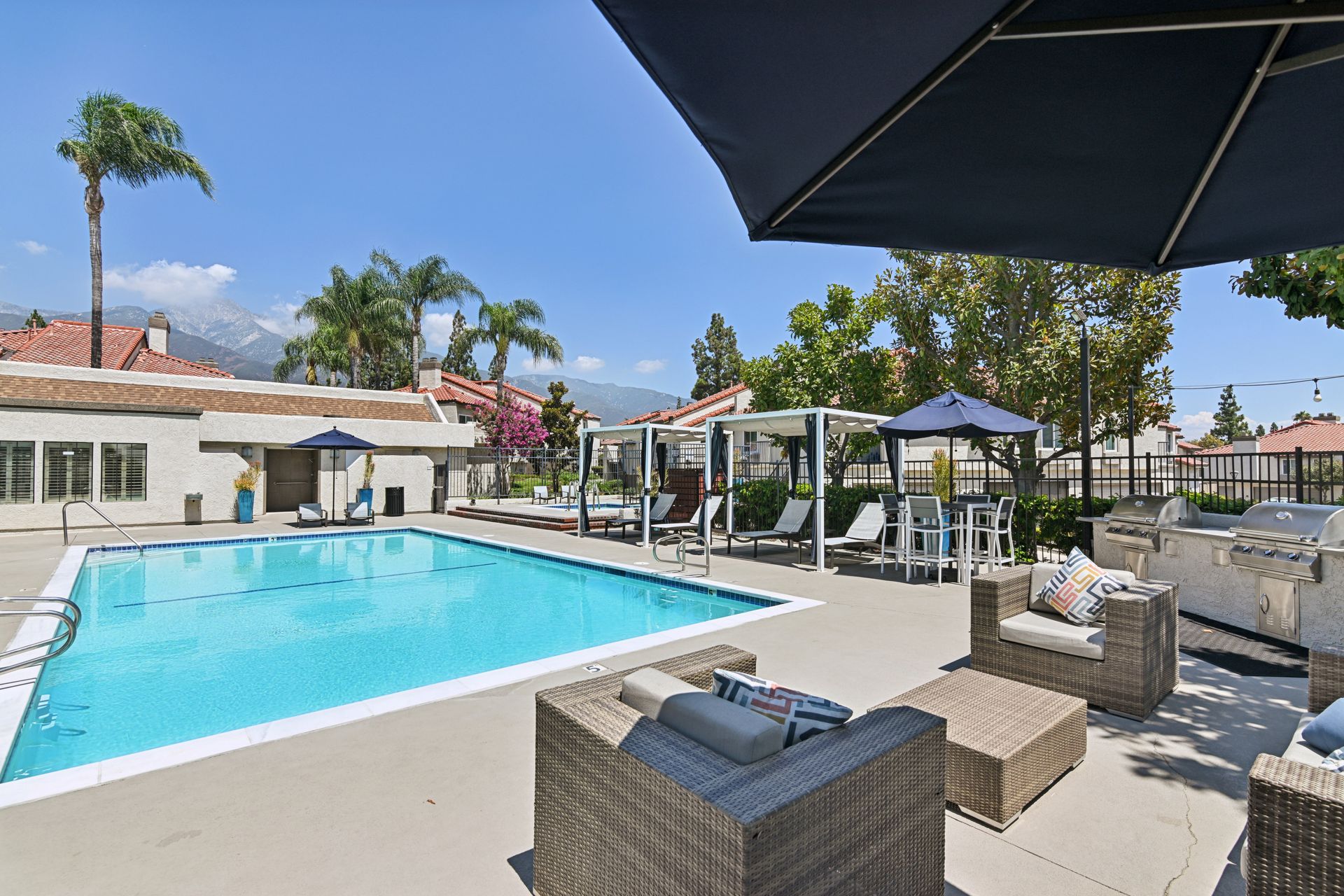 Outdoor pool with lounge chairs and umbrellas on a sunny patio, with nearby buildings and trees.