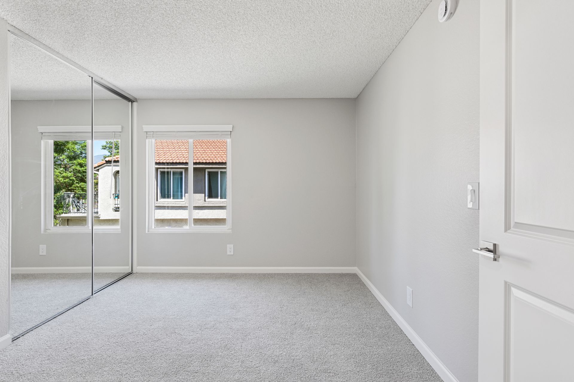 Empty white bedroom with mirrored closet doors and two windows overlooking a building