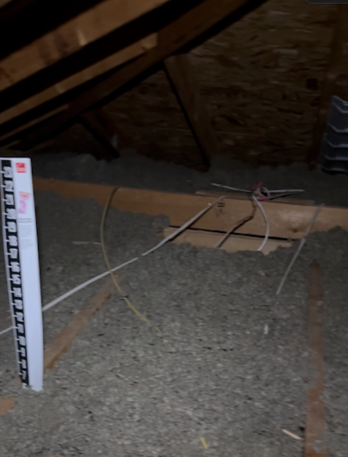 Attic insulation with exposed wires and a ruler standing upright near the rafters
